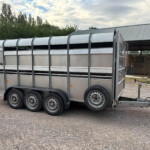 Long silver livestock trailer with curved top, mounted on six wheels, parked on a gravel lot beside a barn.