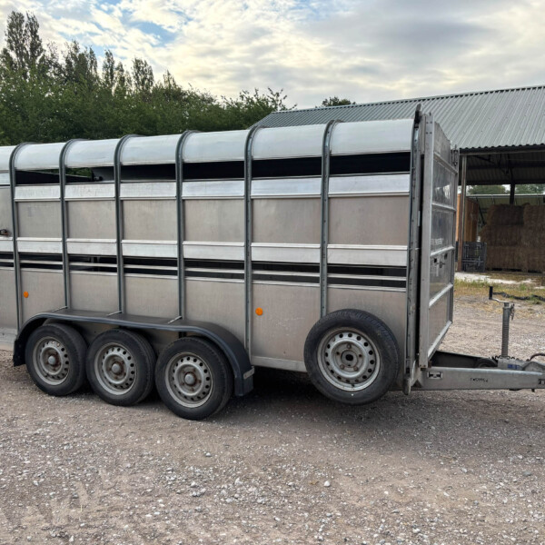 Long silver livestock trailer with curved top, mounted on six wheels, parked on a gravel lot beside a barn.