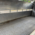 Empty metal trailer interior with diamond-plate floor, riveted walls, and open window slats showing greenery outside.