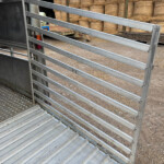 Open livestock trailer showing metal rails and ramp, with stacked hay bales in a farmyard background.
