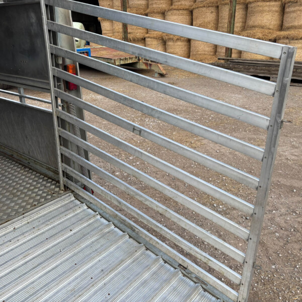 Open livestock trailer showing metal rails and ramp, with stacked hay bales in a farmyard background.