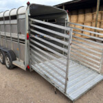 Metal livestock trailer with its ramp lowered, parked on a gravel lot beside a hay-bale storage area. A red truck is partially visible to the left.