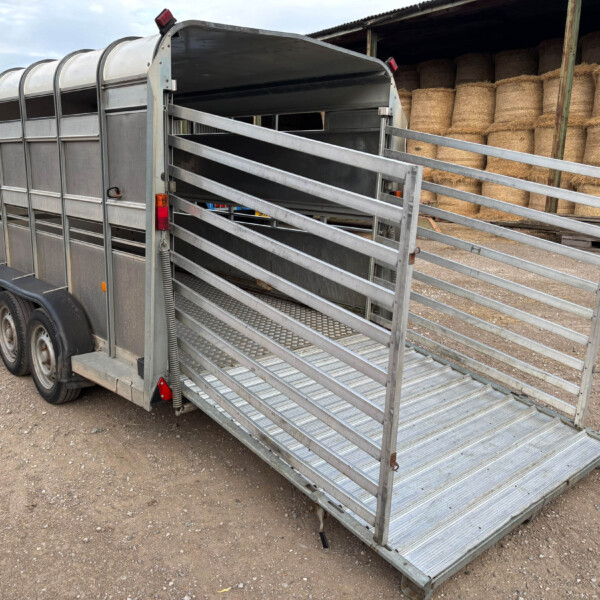 Metal livestock trailer with its ramp lowered, parked on a gravel lot beside a hay-bale storage area. A red truck is partially visible to the left.