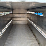 Interior of a metal trailer with diamond-plate floor, side rails, and narrow windows along the top, empty and worn.