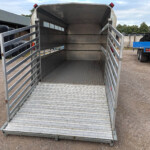 Open metal livestock trailer with rear ramp down, empty interior, in a gravel lot next to stacked tires and a blue trailer.