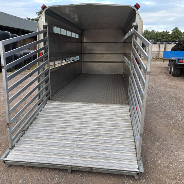 Open metal livestock trailer with rear ramp down, empty interior, in a gravel lot next to stacked tires and a blue trailer.