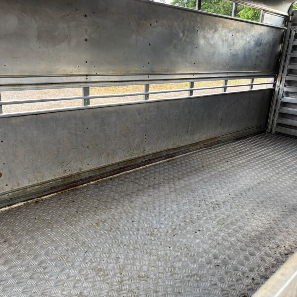 Empty metal trailer interior with diamond-plate floor, riveted walls, and open window slats showing greenery outside.