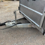 Rear view of a metal trailer with a vertical hand-crank hitch jack and safety chains on a gravel lot, ready to be hitched to a vehicle