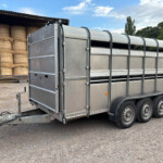 Metal livestock trailer hitched to a truck on a gravel lot with stacked round hay bales inside a barn behind it.