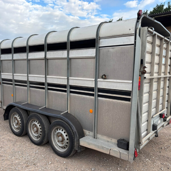 Metal livestock trailer on a gravel lot, with stacked hay bales and a red vehicle in the background.