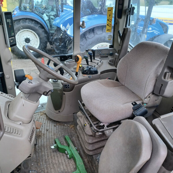 Inside a tractor cab showing the steering wheel, multiple control levers, and a dusty seat; a blue tractor is visible through the window outside.