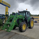 Green John Deere tractor with a large front loader parked outside a dealership under a cloudy sky.