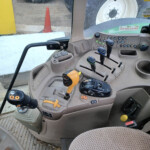 Interior of a tractor cab with multiple control levers, switches, and a steering column visible; windshield shows a yellow post and orange cone outside.