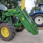 Close-up of green farming machinery with hydraulic arms attached to a tractor, yellow wheels, and other tractors in background.