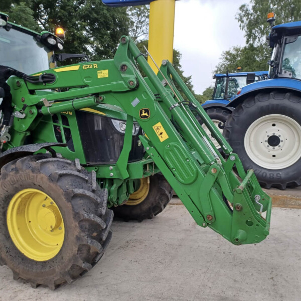 Close-up of green farming machinery with hydraulic arms attached to a tractor, yellow wheels, and other tractors in background.