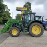 Green New Holland agricultural tractor with a front loader attachment and large yellow wheels parked on a paved lot under a cloudy sky, with trees in the background.