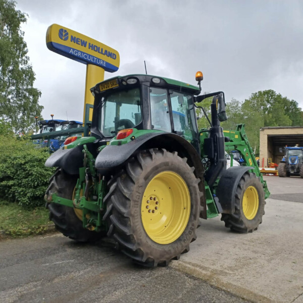 Green and yellow New Holland tractor in foreground with large rear tires, outdoor lot, sign reading 'New Holland Agriculture' in background on cloudy day.