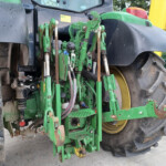 Rear view of a green farming implement attached to a tractor with large muddy tires, yellow wheel rims, and hitch hardware visible.