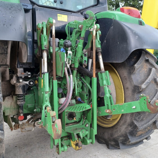 Rear view of a green farming implement attached to a tractor with large muddy tires, yellow wheel rims, and hitch hardware visible.