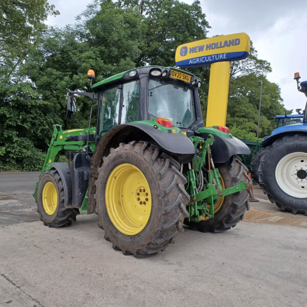 Two tractors parked outdoors in a lot, a green John Deere in front and a blue tractor to the right beneath a New Holland Agriculture sign used at a dealership.
