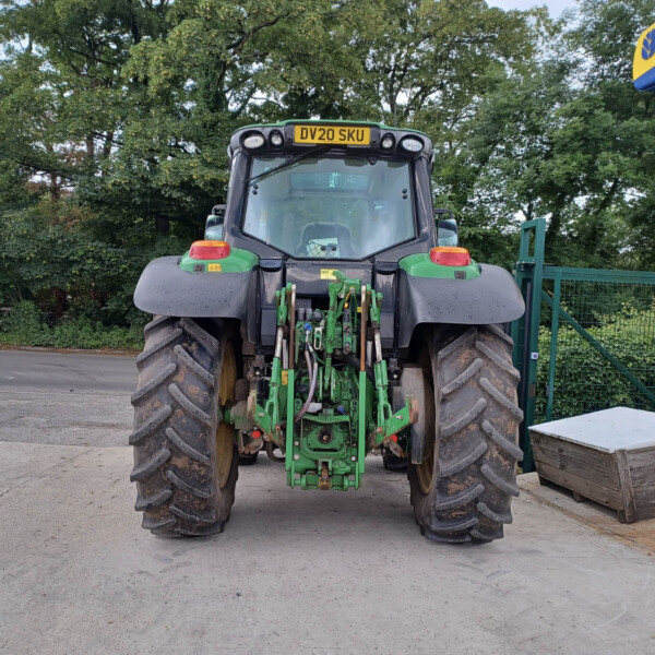Rear view of a large green tractor with mounted hydraulic equipment, license plate DV20 SKU, on a concrete lot with trees in the background.