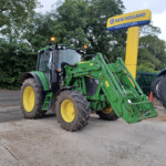 Two green John Deere tractors with big yellow wheels parked on a concrete lot near a New Holland Agriculture sign in the background