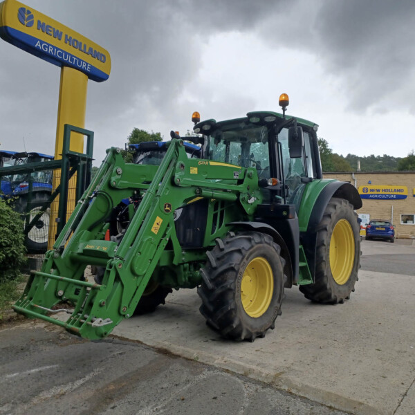 Green John Deere tractor with a large front loader parked outside a dealership under a cloudy sky.