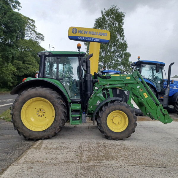 Two green New Holland tractors with yellow wheels parked in a lot under a yellow New Holland Agriculture sign.