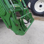 Green agricultural implement frame with hydraulic cylinders and hoses, large white tractor tire in background on a concrete surface