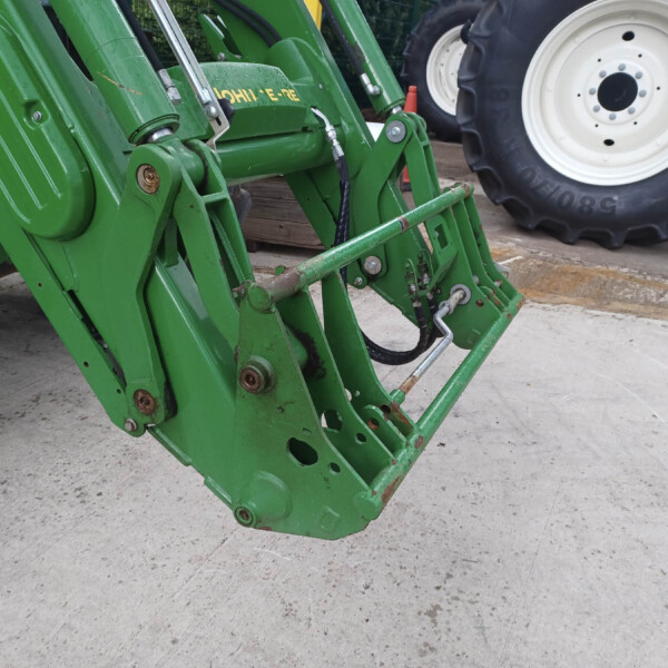 Green agricultural implement frame with hydraulic cylinders and hoses, large white tractor tire in background on a concrete surface