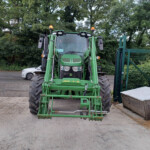 Front view of a green John Deere tractor with front loader arms, parked outdoors near a fence and leafy trees.