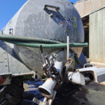 Large galvanized tanker trailer with green hoses draped along its side, parked beside a wooden shed in a yard