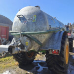 Silver cylindrical tanker trailer with green hoses attached, parked in an industrial yard beside a red Baileys trailer and metal buildings.