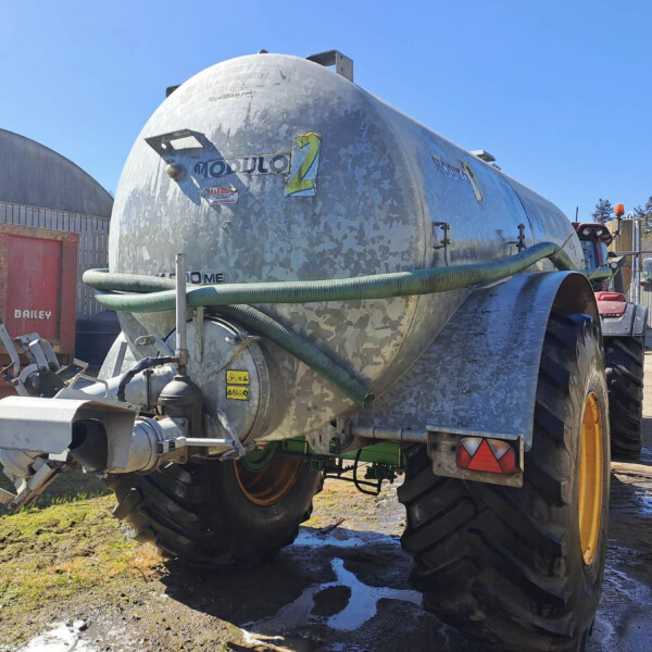 Silver cylindrical tanker trailer with green hoses attached, parked in an industrial yard beside a red Baileys trailer and metal buildings.