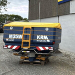 Rear view of a blue trailer with a bright yellow tarp, strapped down, sitting on wooden pallets in a gravel yard; farm equipment visible in the background.