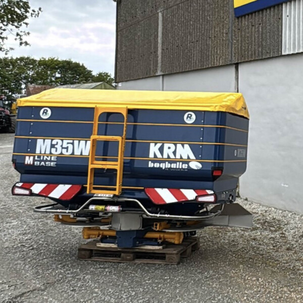 Rear view of a blue trailer with a bright yellow tarp, strapped down, sitting on wooden pallets in a gravel yard; farm equipment visible in the background.