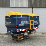 Blue and yellow mobile elevated work platform (MEWP) parked on pallets outdoors against a gray concrete wall, with a protective canopy.