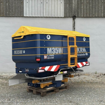 Blue and yellow mobile elevated work platform (MEWP) parked on pallets outdoors against a gray concrete wall, with a protective canopy.