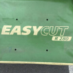 Close-up of a green metal surface with a beige 'EASYCUT' logo and 'R280' label; small circular holes and a light-colored rod along the bottom.