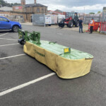 Green Krone mower attachment in a parking lot foreground, with a blue pickup and industrial yard in the background.