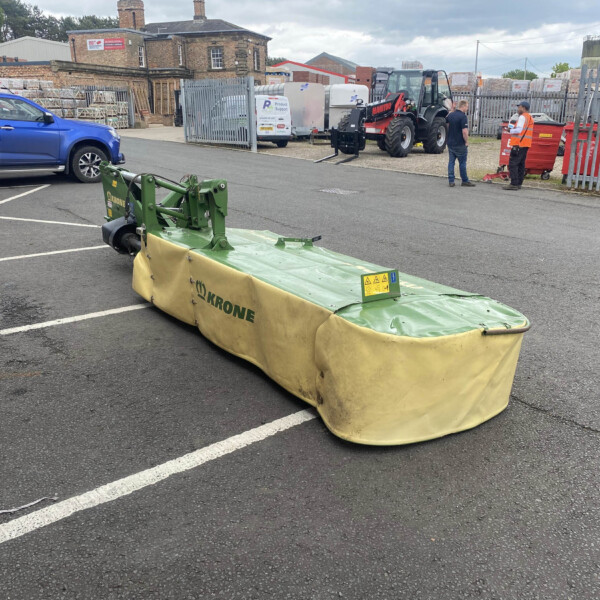 Green Krone mower attachment in a parking lot foreground, with a blue pickup and industrial yard in the background.