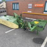 Green agricultural mower attachment parked in a paved lot beside a brick building with Honda and Kubota signs on the wall.