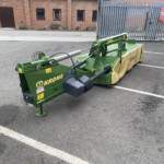 Green Krone agricultural mower attachment in a parking lot, with a brick building and metal fence in the background.