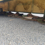 Underneath a yellow, rust-streaked industrial deck with circular rusted weights and green moss, resting on a pebbled surface