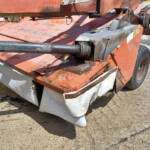 Close-up of an orange industrial machine showing a rusty hydraulic drive shaft and worn panels near a large rear wheel on a dirt surface