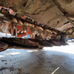 Underbelly of a damaged boat hull with numerous white propeller blades exposed amid dirt and debris in a salvage yard, with parked trucks in the background.