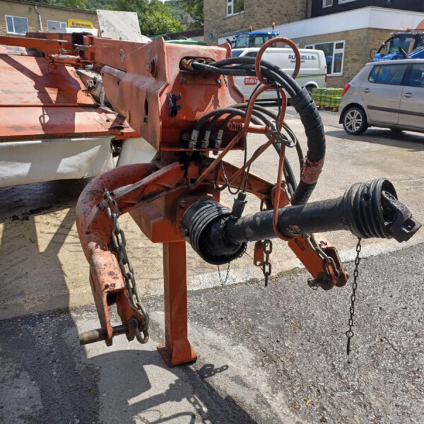 Industrial orange hydraulic machine with hoses and chains in a dusty yard near a car and construction equipment.