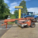 New Holland Agriculture sign with a yellow pole and blue tractors on a trailer in a parking lot under a blue sky and green trees.