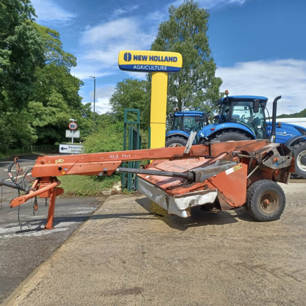 New Holland Agriculture sign with a yellow pole and blue tractors on a trailer in a parking lot under a blue sky and green trees.