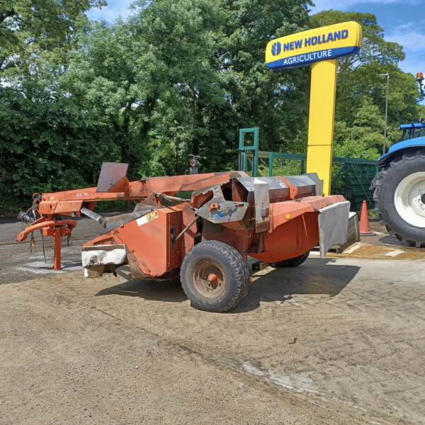 Orange agricultural trailer with metal framing, parked in a lot beside a blue New Holland tractor, with green trees in the background.
